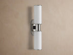 Harlin Double Sconce with two glowing vertical milk glass shades and polished nickel accents, mounted on a textured brown wall.