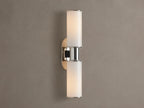 Harlin Double Sconce with two glowing vertical milk glass shades and polished nickel accents, mounted on a textured brown wall.