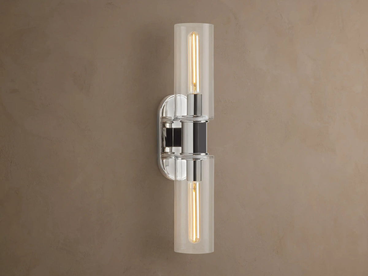 Vertical Harlin Double Sconce with polished nickel finish and two clear glass cloches revealing illuminated Edison bulbs on a brown wall.