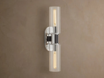 Vertical Harlin Double Sconce with polished nickel finish and two clear glass cloches revealing illuminated Edison bulbs on a brown wall.