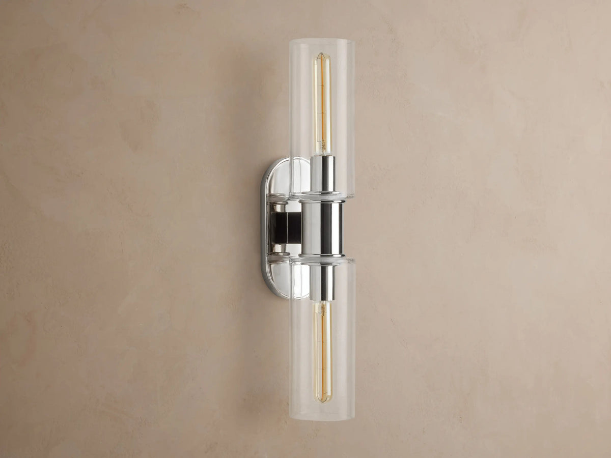 Vertical Harlin Double Sconce with polished nickel finish and two clear glass cloches revealing illuminated Edison bulbs on a brown wall.