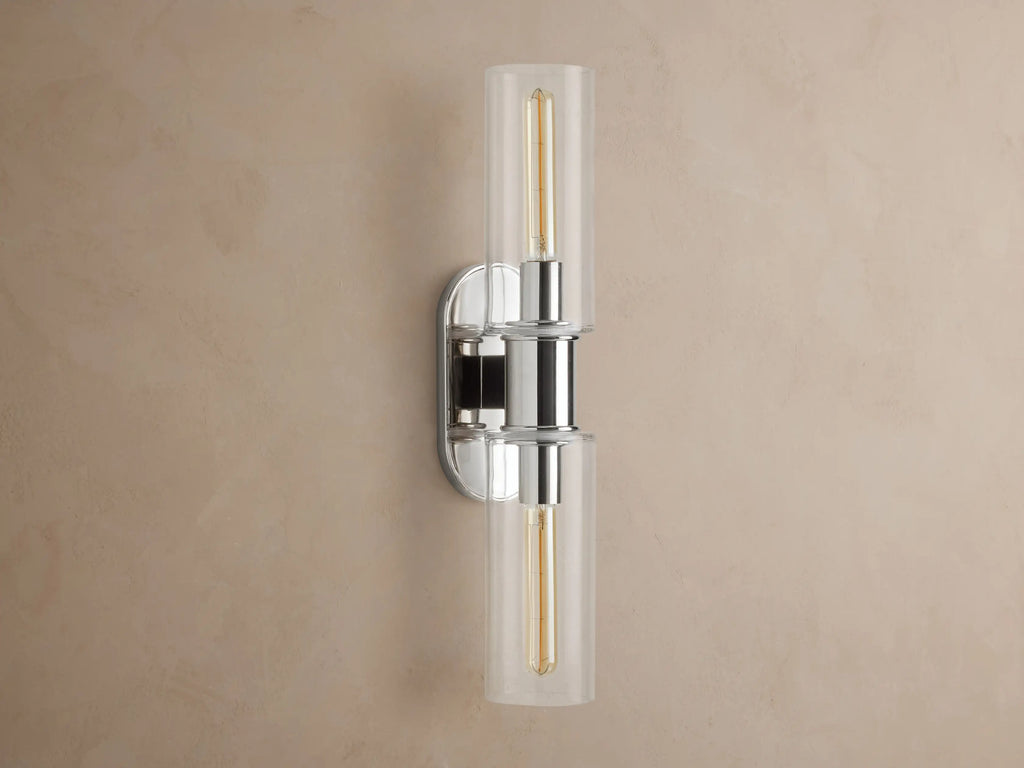 Vertical Harlin Double Sconce with polished nickel finish and two clear glass cloches revealing illuminated Edison bulbs on a brown wall.