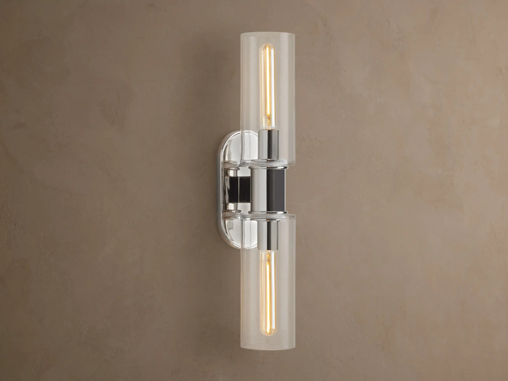 Vertical Harlin Double Sconce with polished nickel finish and two clear glass cloches revealing illuminated Edison bulbs on a brown wall.