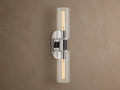 Vertical Harlin Double Sconce with polished nickel finish and two clear glass cloches revealing illuminated Edison bulbs on a brown wall.