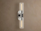 Vertical Harlin Double Sconce with polished nickel finish and two clear glass cloches revealing illuminated Edison bulbs on a brown wall.