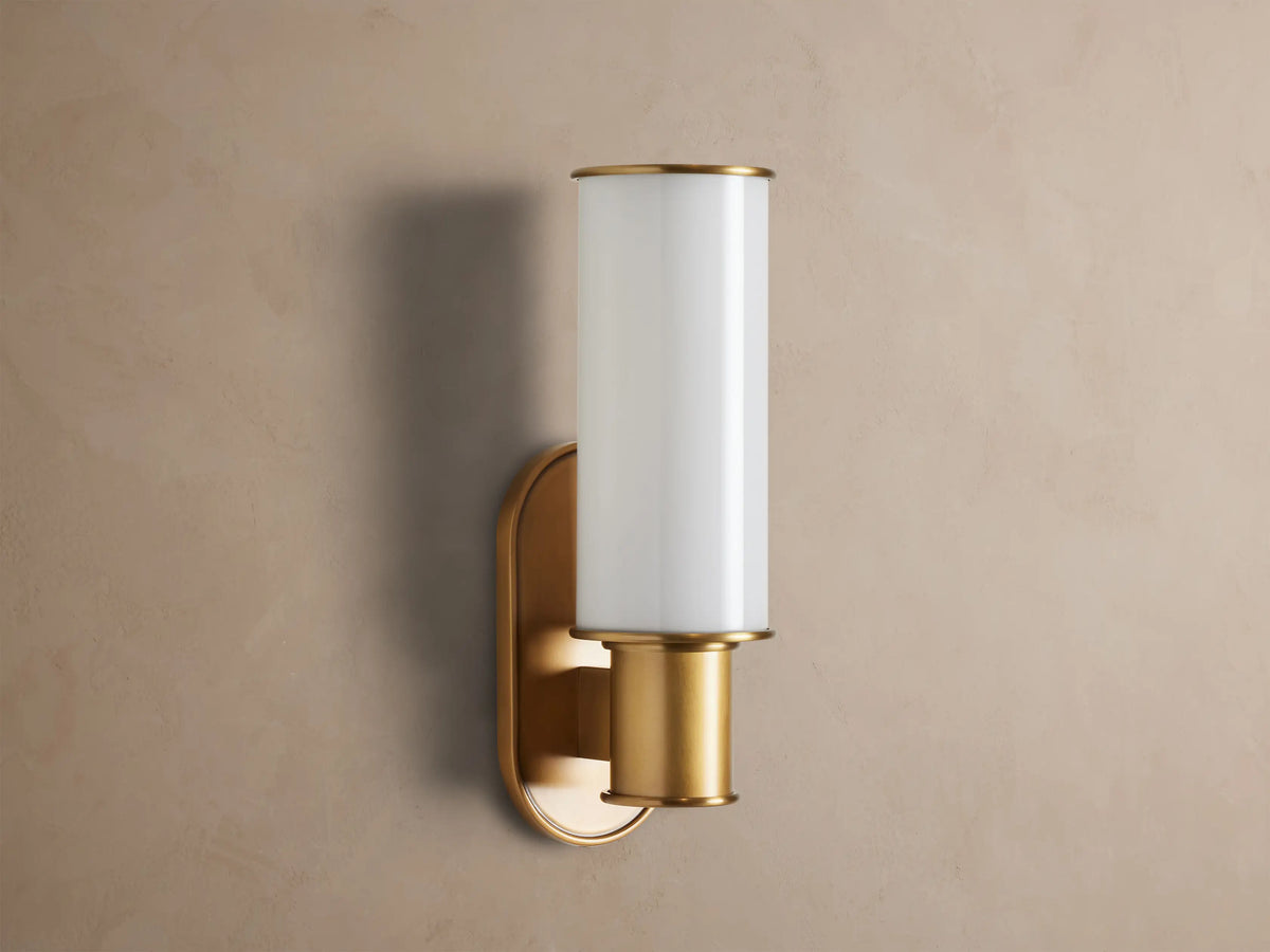 Harlin Sconce with a vertical milk glass cylindrical shade and burnished brass base, illuminated on a textured brown wall.