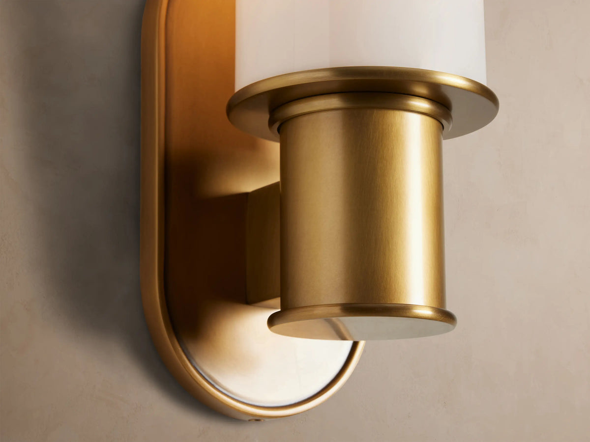 Harlin Sconce with burnished brass oval backplate and a white milk glass cloche, mounted vertically on a beige wall.