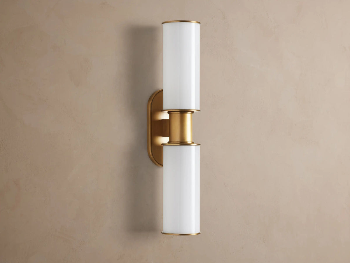 Harlin Double Sconce mounted vertically on a wall. It has two glowing milk glass cylinders separated by a burnished brass stem & oval backplate.