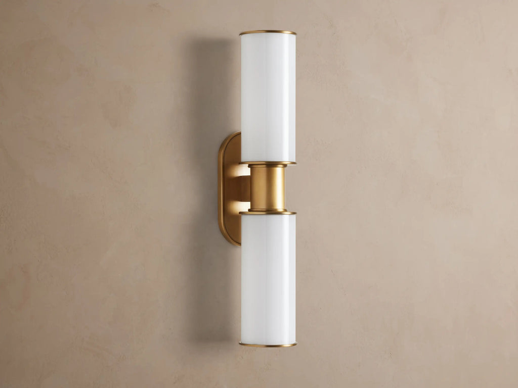 Harlin Double Sconce mounted vertically on a wall. It has two glowing milk glass cylinders separated by a burnished brass stem & oval backplate.