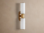 Harlin Double Sconce mounted vertically on a wall. It has two glowing milk glass cylinders separated by a burnished brass stem & oval backplate.