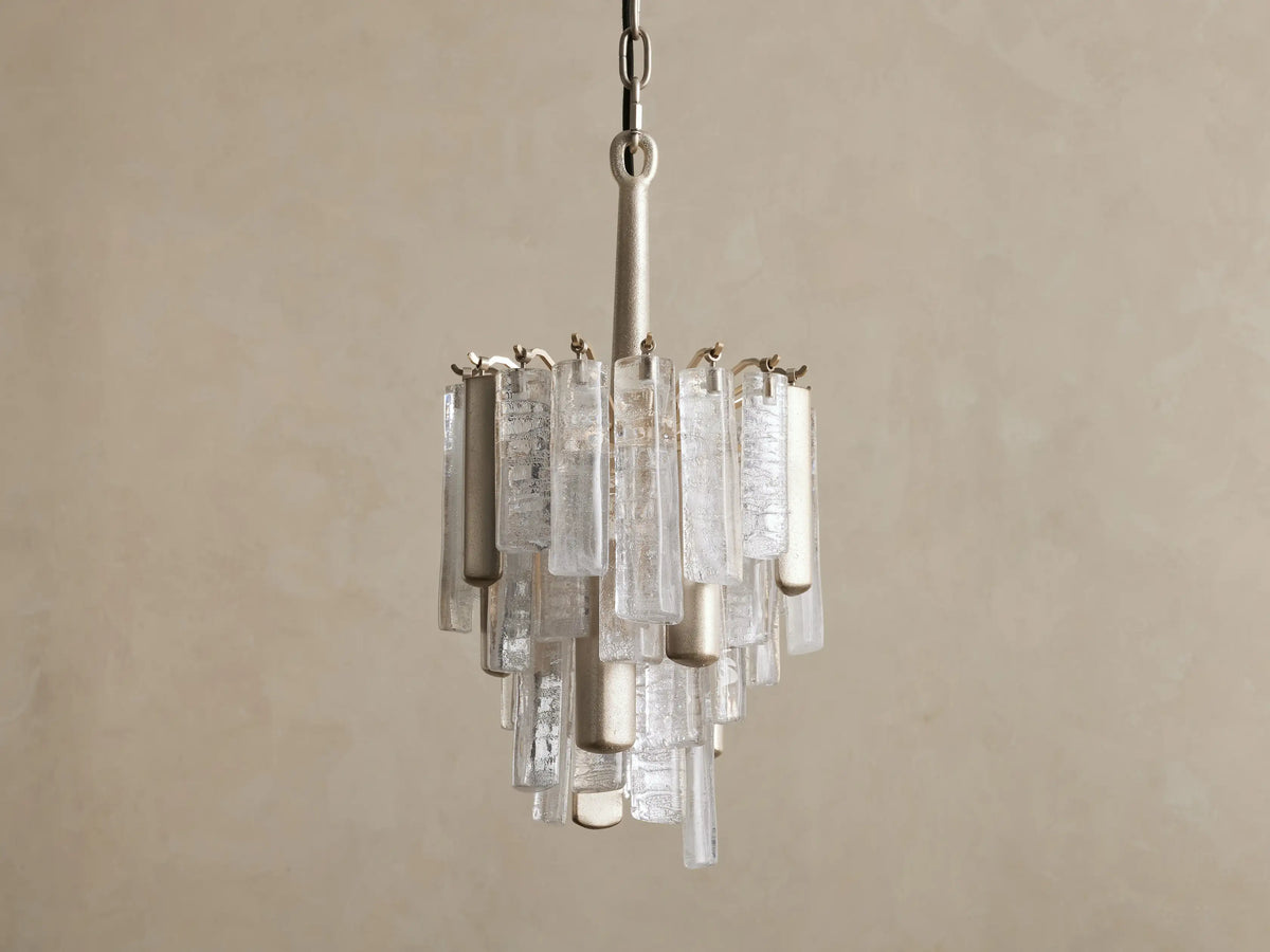 Neve Pendant: Brass-finished pendant with cascading clear textured cast glass elements glowing warmly against a taupe wall.