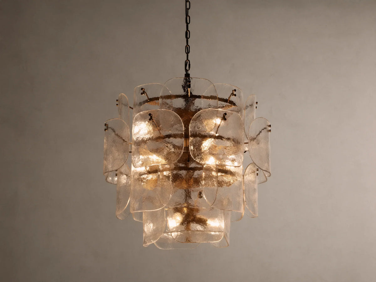 Blaire Round Chandelier with layered, curved clear glass panels creating a textured effect. Lit with warm light, hung by a brass-finished chain.