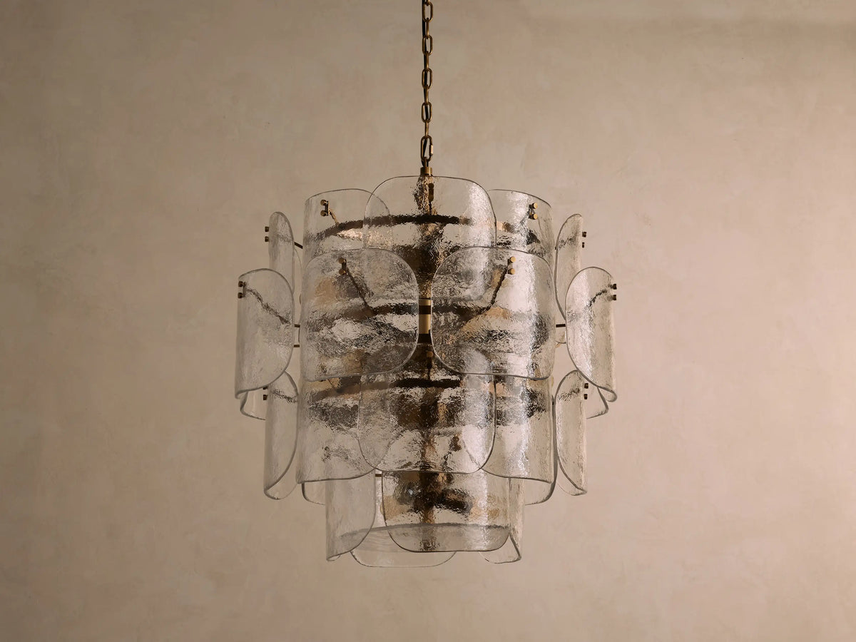 Blaire Round Chandelier with layered, curved clear glass panels creating a textured effect. Lit with warm light, hung by a brass-finished chain.