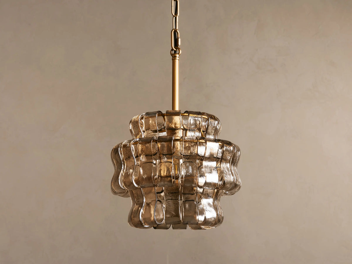 Ghiaccio Pendant with tiered curved glass design, glowing softly. Brass stem and hanging chain. Warm, ambient lighting effect.