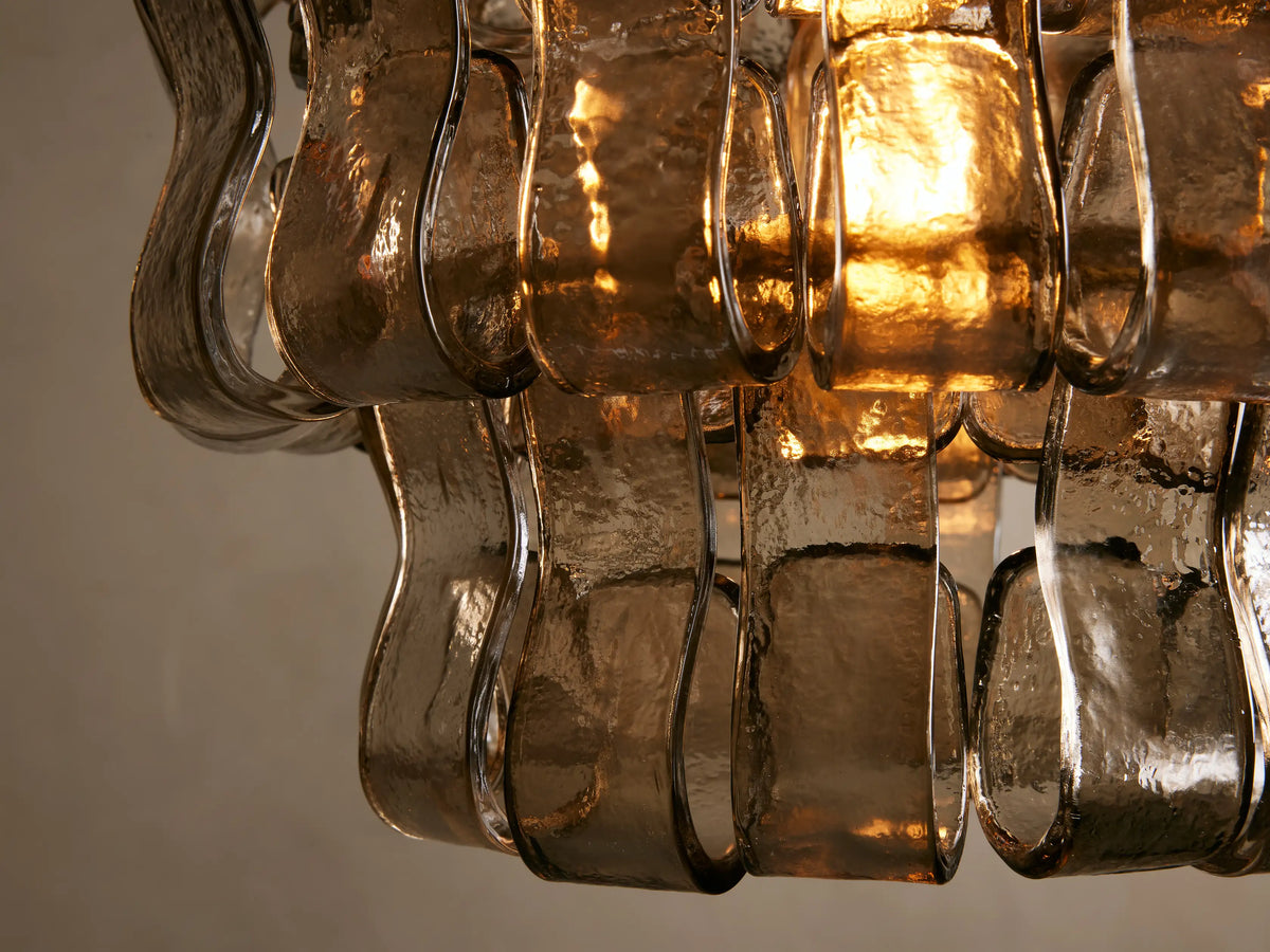 Curved glass pieces of the Ghiaccio Pendant, showcasing textured amber-toned glass and steel accents with warm light reflections.