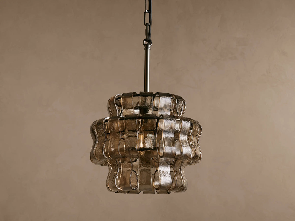 Ghiaccio Pendant with wavy curved glass panels illuminated from within, hanging on a black steel chain against a brown backdrop.