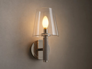 Monroe Sconce in polished nickel finish with a clear glass shade, emitting warm light mounted on a beige wall beside a mirror.