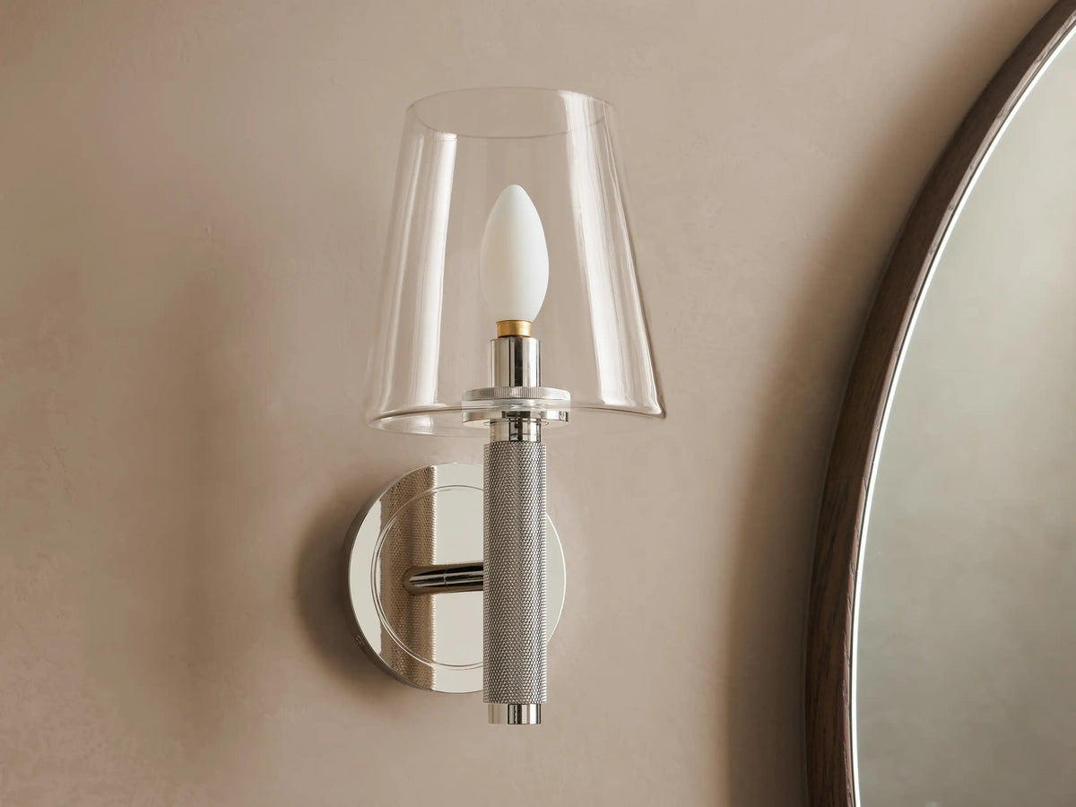 Monroe Sconce in polished nickel finish with a clear glass shade, emitting warm light mounted on a beige wall beside a mirror.