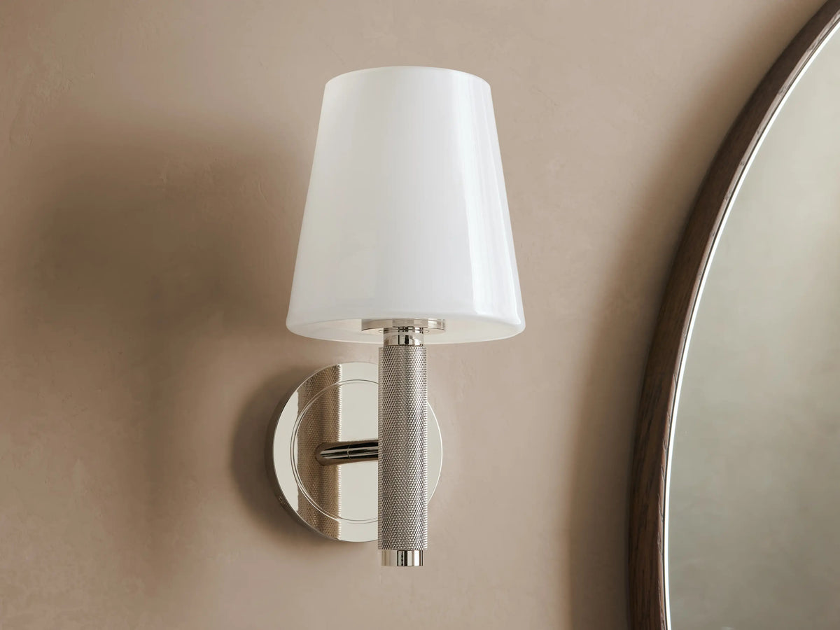 Monroe Sconce in polished nickel finish with a lit white glass shade, mounted on a beige wall beside a mirror.