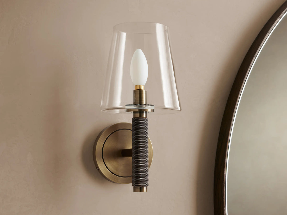 Monroe Sconce with antique brass finish and clear glass shade, glowing warmly on a textured wall next to a dark mirror.