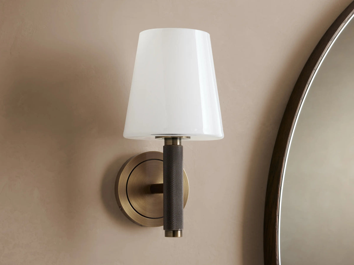 Monroe Sconce with antique brass finish, round wall mount, and soft glowing milk glass shade, shown beside mirror.