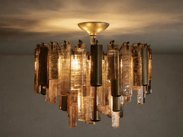 Neve Flush Mount, a round ceiling light with textured bronze cast glass rods & brass-finished aluminum cylinders, lit with a warm glow.