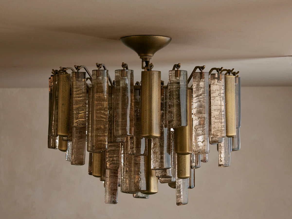 Neve Flush Mount, a round ceiling light with textured bronze cast glass rods & brass-finished aluminum cylinders, lit with a warm glow.