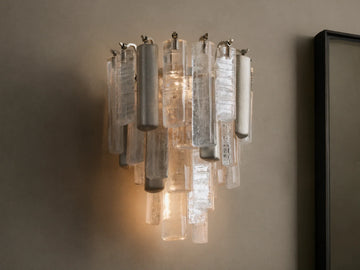 Neve Sconce: Wall-mounted light with clear textured glass and champagne-finished aluminum elements, glowing warmly.