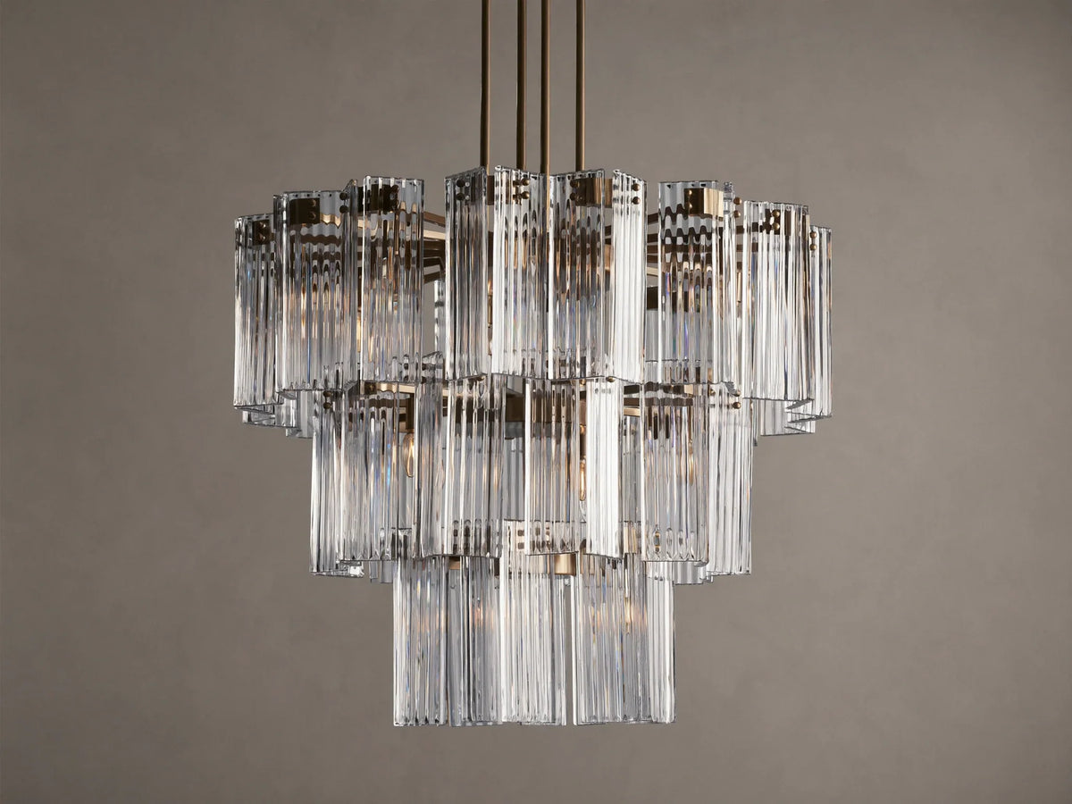 Delsie Round Chandelier, multi-tiered with clear ribbed glass rods and burnished brass frame, hanging against a brown wall.