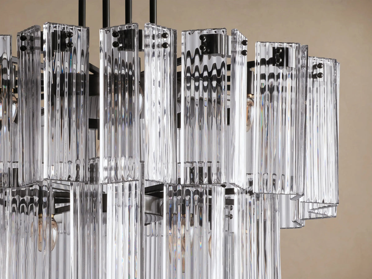 Delsie Round Chandelier: tiered, clear fluted glass rods with a dark metal frame, hanging against a light brown background.