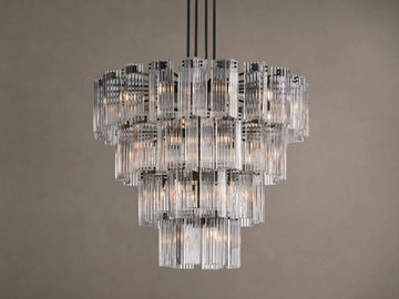 Delsie Round Chandelier with illuminated tiers of clear, fluted glass rods and a dark metal frame hanging against a taupe wall.