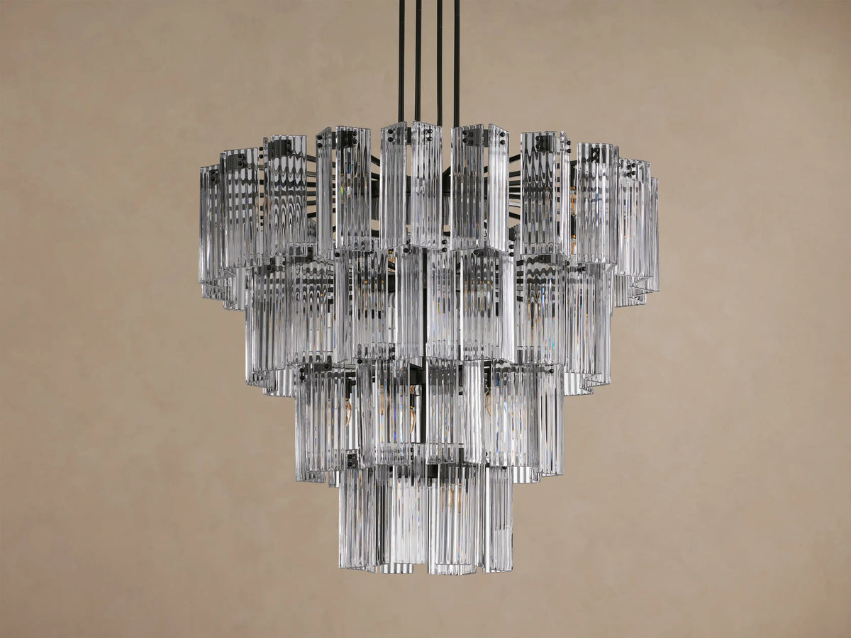 Delsie Round Chandelier with illuminated tiers of clear, fluted glass rods and a dark metal frame hanging against a taupe wall.