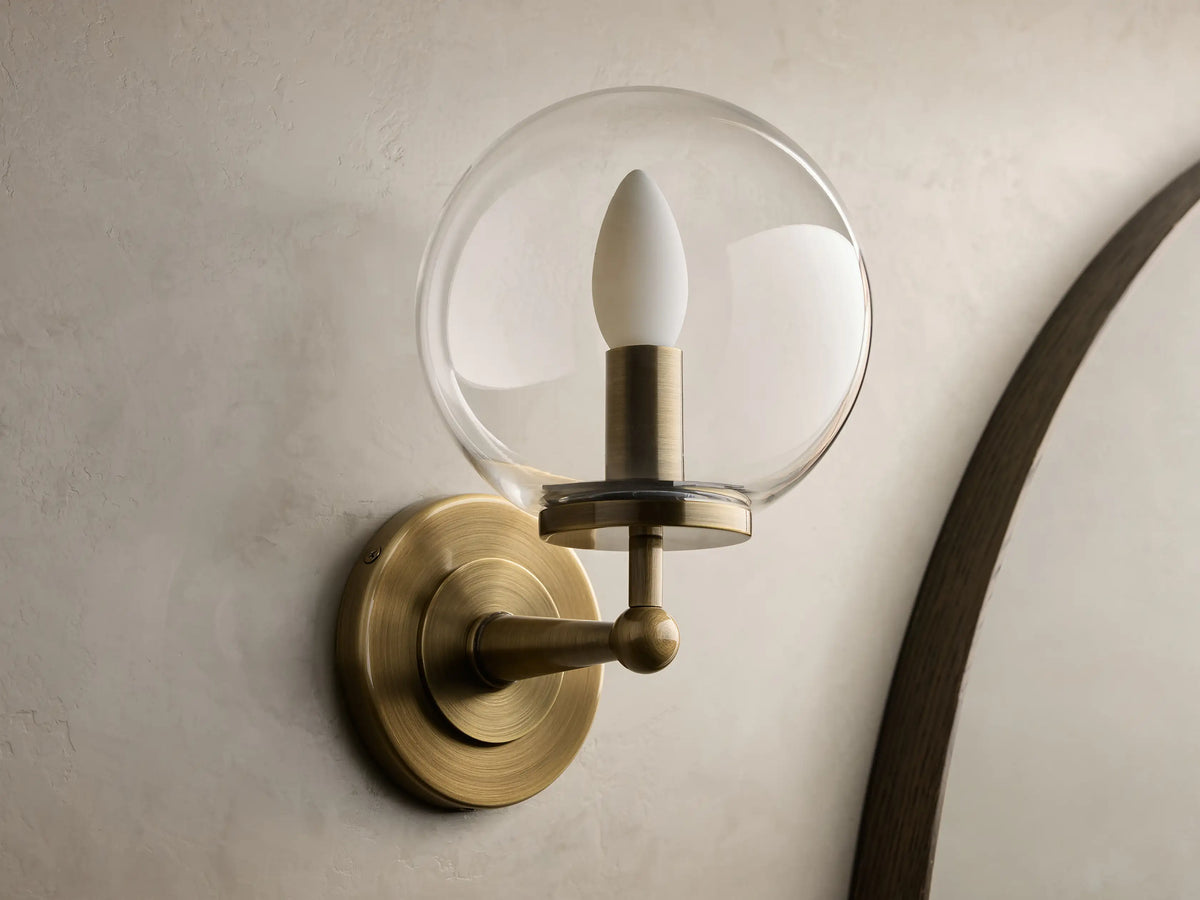 Front view of an antique brass Fulton Sconce, featuring a round backplate, arm, and clear glass globe enclosing a white bulb.
