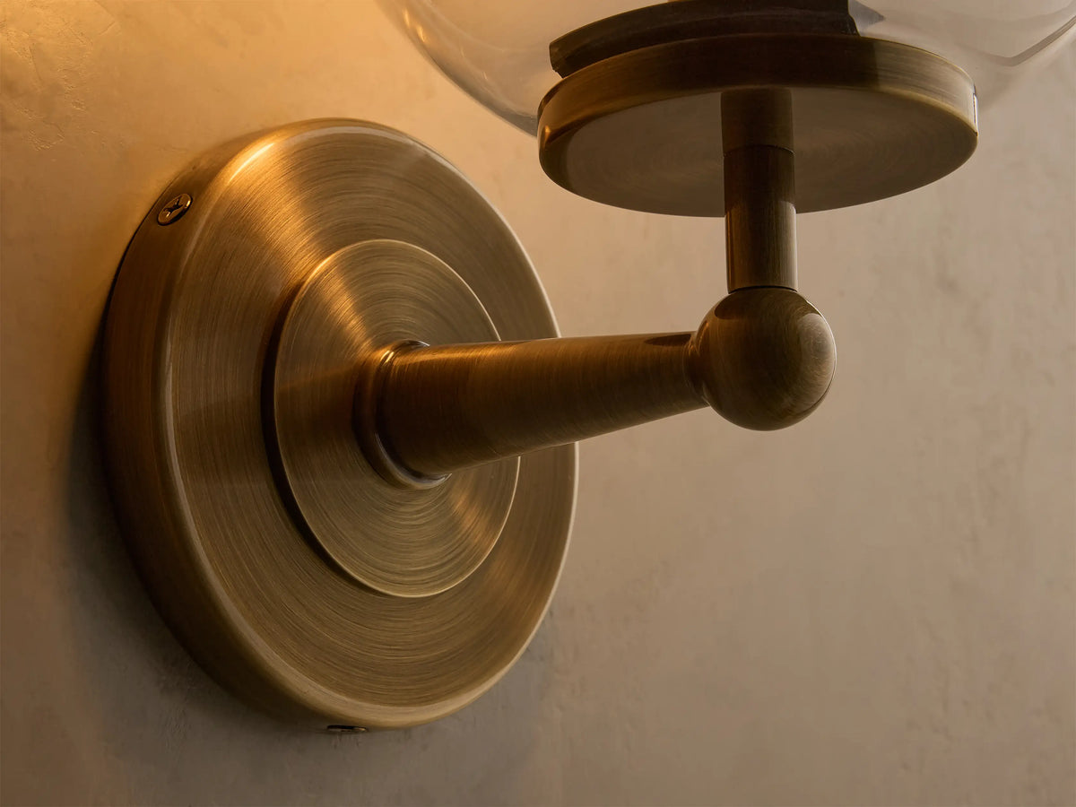 Front view of the antique brass Fulton Sconce featuring a lit bulb within a clear mouth-blown glass globe.