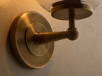 Front view of the antique brass Fulton Sconce featuring a lit bulb within a clear mouth-blown glass globe.
