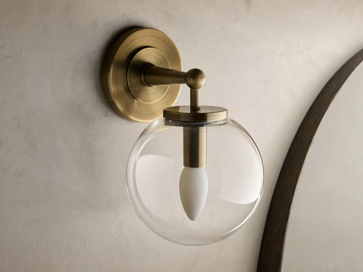 Close-up of the antique brass Fulton Sconce, highlighting its brushed finish, round backplate, and part of a clear glass globe.