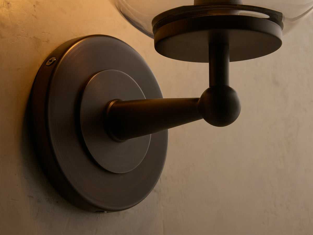 Close-up of the antique bronze Fulton Sconce with a clear glass globe and an illuminated bulb, mounted on a light wall.