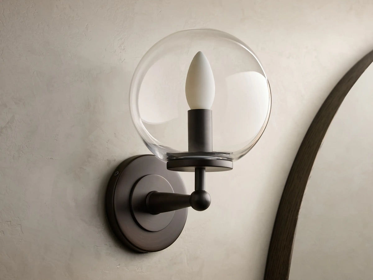 Close-up of the antique bronze Fulton Sconce with a clear glass globe, mounted on a textured wall.
