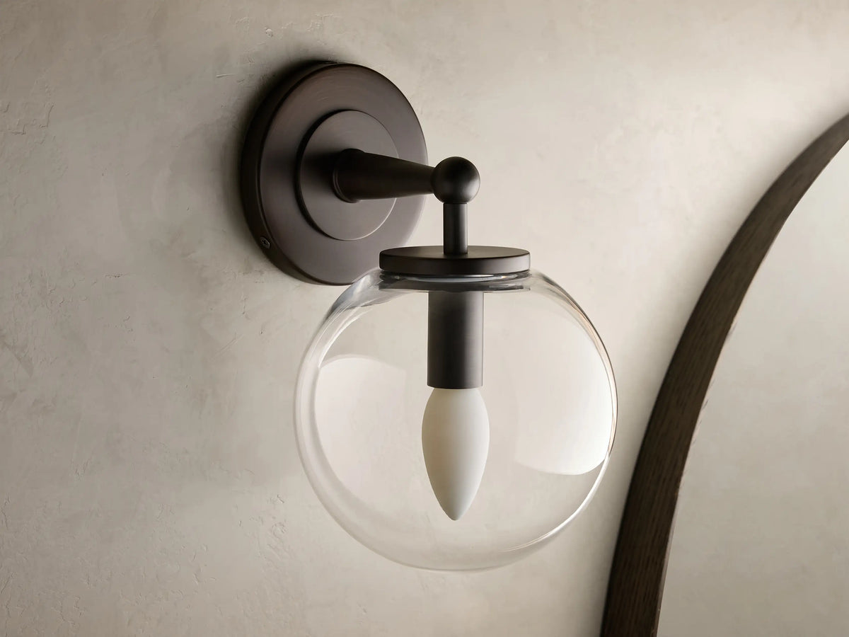 Close-up of the antique bronze Fulton Sconce with a clear glass globe housing a white bulb, mounted on a textured wall.