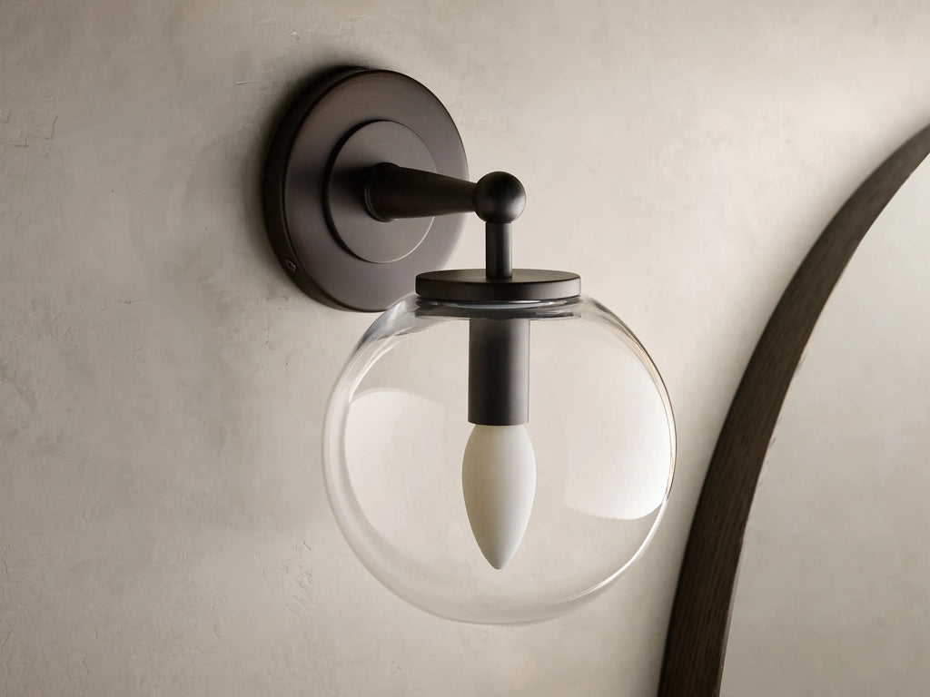 Close-up of the antique bronze Fulton Sconce with a clear glass globe housing a white bulb, mounted on a textured wall.