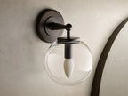 Close-up of the antique bronze Fulton Sconce with a clear glass globe housing a white bulb, mounted on a textured wall.
