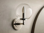 Front view of Polished Nickel Fulton Sconce with a clear glass globe and a white candle-shaped bulb on a gray wall.