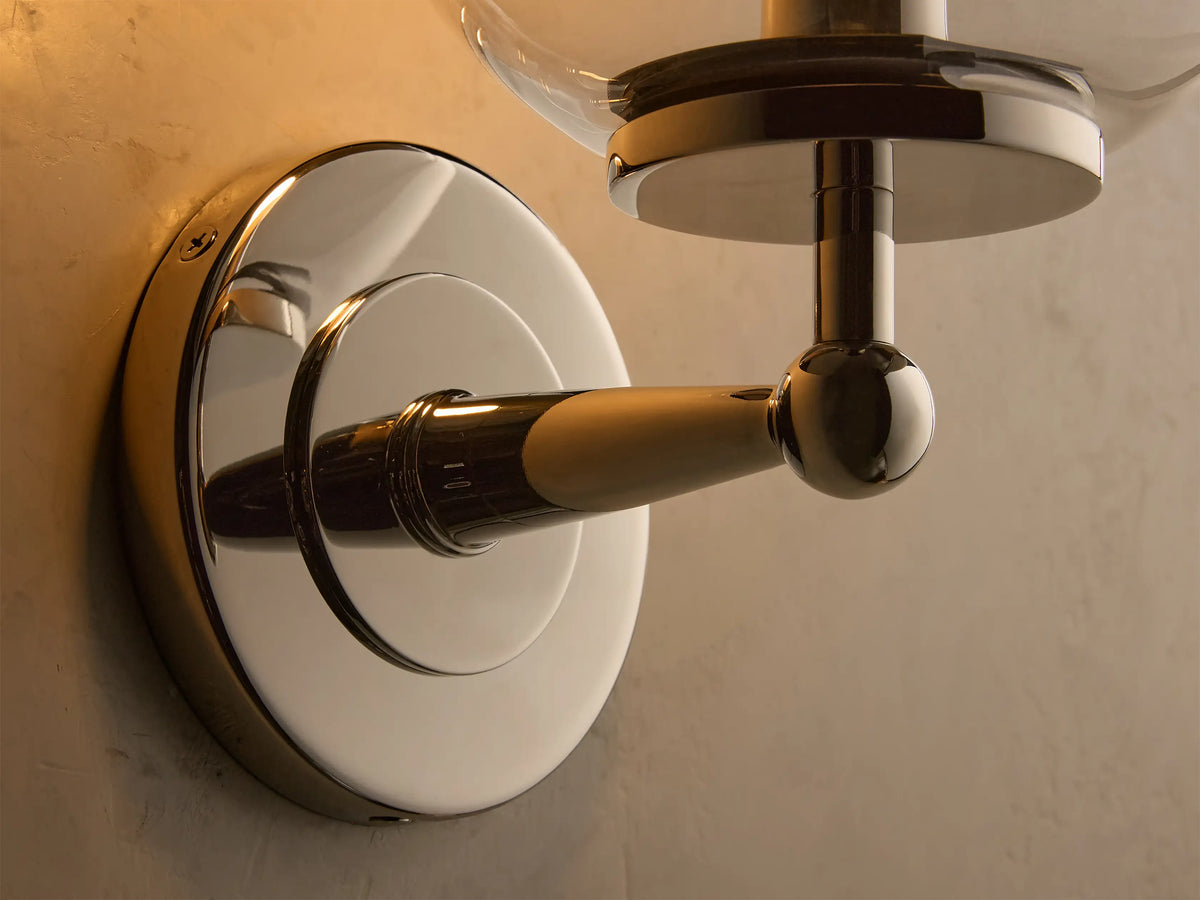 Front view of polished nickel Fulton Sconce with a clear glass globe and illuminated bulb on a textured beige wall.