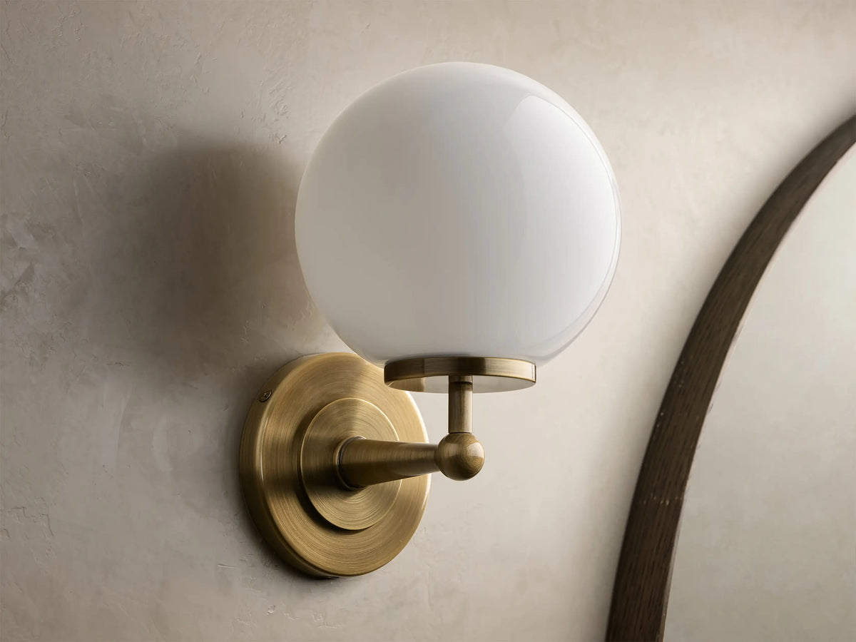 Front view of an antique brass Fulton Sconce with a glowing, round milk glass globe, mounted on a textured wall.