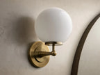 Front view of an antique brass Fulton Sconce with a glowing, round milk glass globe, mounted on a textured wall.
