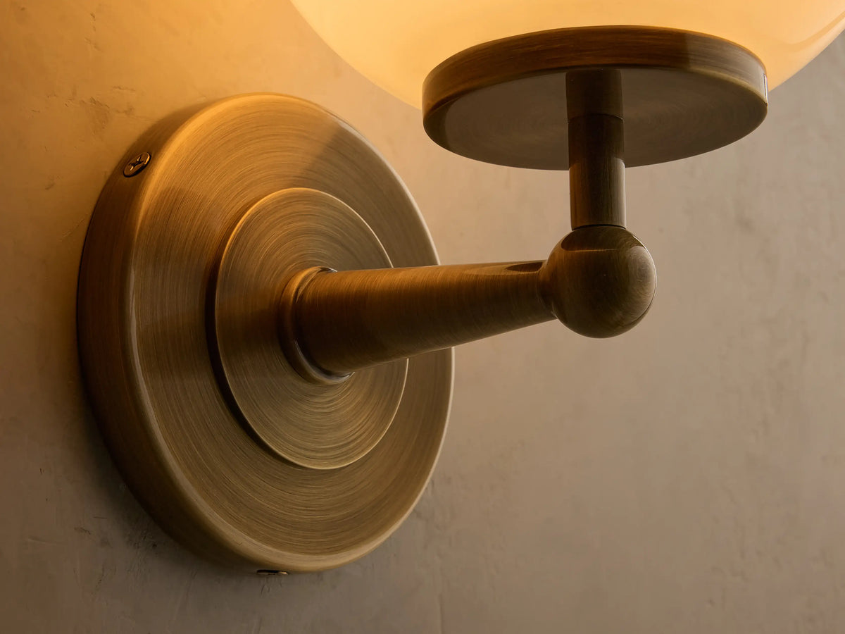 Front view of an antique brass Fulton Sconce featuring a spherical milk glass globe, mounted on a light textured wall.