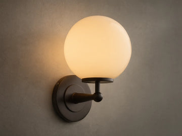 Front view of the Antique Bronze Fulton Sconce, featuring a lit milk glass spherical globe, mounted on a textured gray wall.
