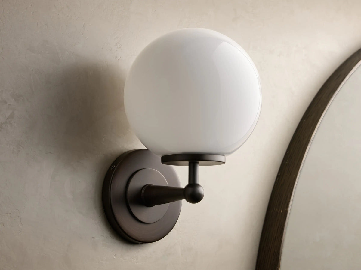 Front view of the Antique Bronze Fulton Sconce, featuring a lit milk glass spherical globe, mounted on a textured gray wall.