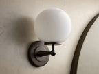 Front view of the Antique Bronze Fulton Sconce, featuring a lit milk glass spherical globe, mounted on a textured gray wall.