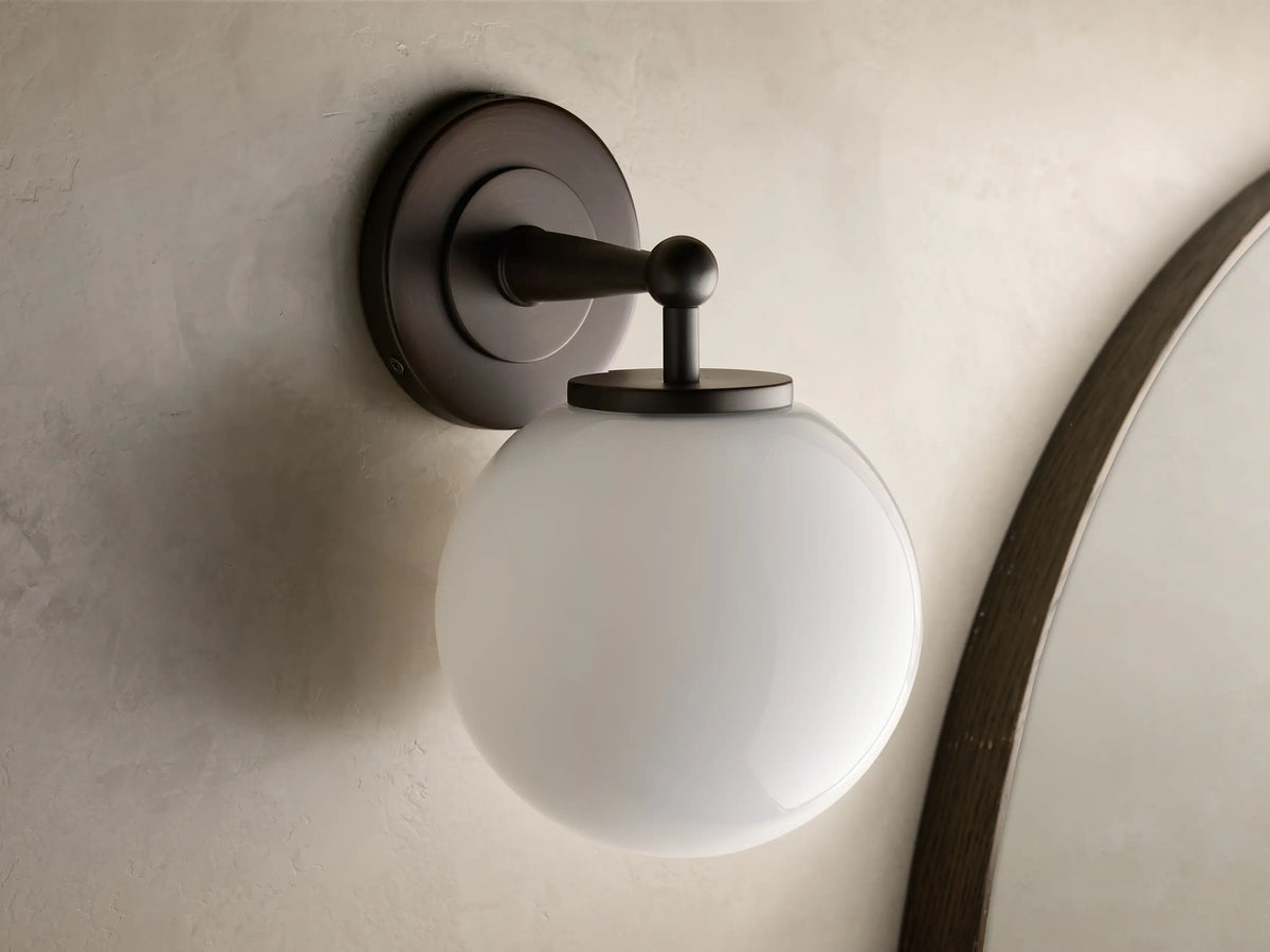 Close-up of the antique bronze Fulton Sconce with a glowing milk glass globe attached to a round backplate.